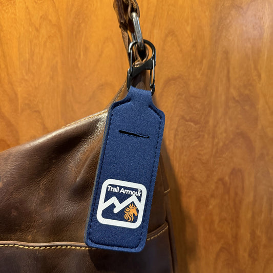 Navy neoprene Trail Armour lip balm holder clipped to a leather bag strap, showing the metal snap clip and the slot in the front